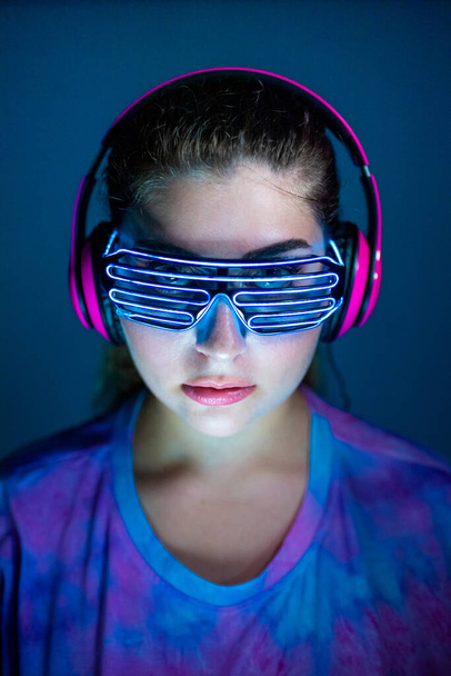 ヘッドフォンを身に着けている若い女性と青の背景に未来的な主導の眼鏡-ネオンのメガネとヘッドフォンを身に着けているサイレントディスコをクラブ孤立した女性-パーティー、未来、技術コンセプト  - 写真・画像