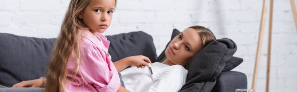 Οριζόντια εικόνα του παιδιού κρατώντας θερμόμετρο δίπλα στην άρρωστη μητέρα και κοιτάζοντας την κάμερα  - Φωτογραφία, εικόνα