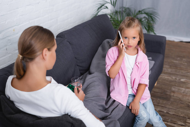 Selective focus of kid talking on smartphone beside mother holding glass of water and pills on couch  - Photo, Image