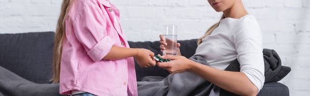 Foto panorámica de niño dando píldoras a la mujer enferma con vaso de agua en el sofá  - Foto, Imagen