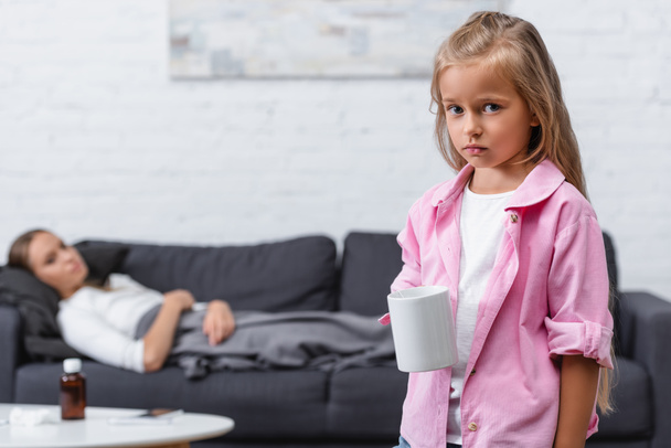 Selective focus of sad girl holding cup with sick woman on couch at background  - Photo, image