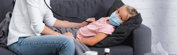 Horizontales Bild des Kindes in medizinischer Maske, das Mutter mit Stethoskop auf Couch betrachtet  - Foto, Bild