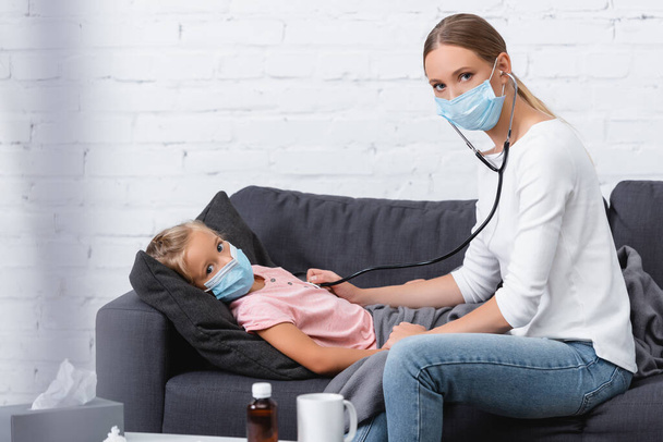 Selective focus of woman in medical mask using stethoscope near sick child on couch  - Foto, Bild