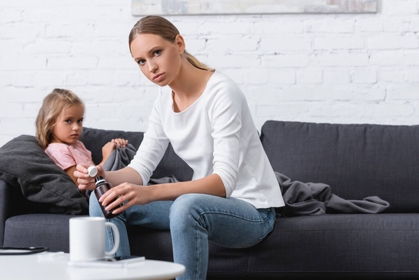 Selektiver Fokus der Frau, die bei Krankheit zu Hause eine Flasche Sirup in der Nähe des Kindes hält  - Foto, Bild