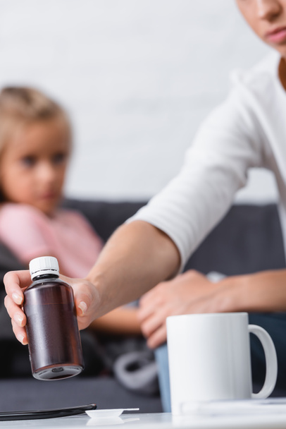 Selective focus of woman taking bottle of syrup near sick child at home  - Fotoğraf, Görsel