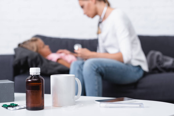 Focus selettivo di pillole, sciroppo e smartphone sul tavolo vicino alla madre seduta accanto al bambino malato a casa  - Foto, immagini