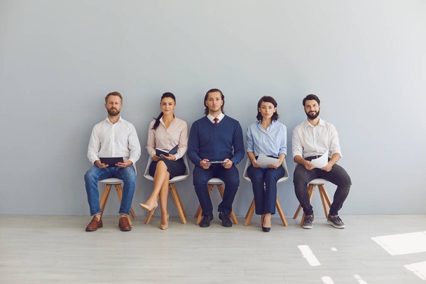 A group of business people are waiting for an interview for a new job. Businessmen and businesswomen sitting on chairs with documents on gray background - Photo, Image