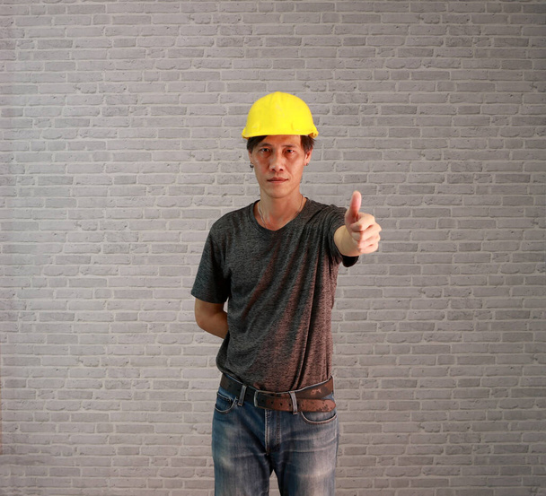 Uomo tecnico ware giallo casco con grigio scuro T-shirt e jeans in denim in piedi e pollici su sfondo grigio modello mattone. - Foto, immagini