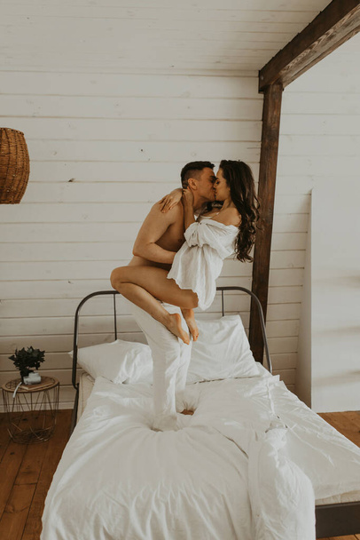 Beautiful young couple in love on the bed in the bedroom. The guy holds the girl in his arms. Kiss each other. - Foto, afbeelding