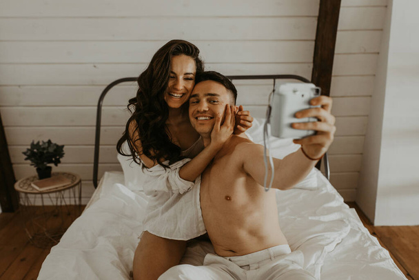  Belo jovem casal apaixonado na cama abraçando rindo e tirando fotos - Foto, Imagem