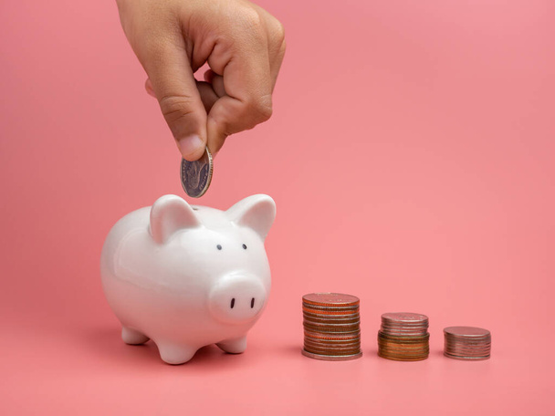 Banco de porquinho branco e moeda e mão humana colocando moeda no banco de porquinho em um fundo rosa. Espaço de cópia para design. - Foto, Imagem
