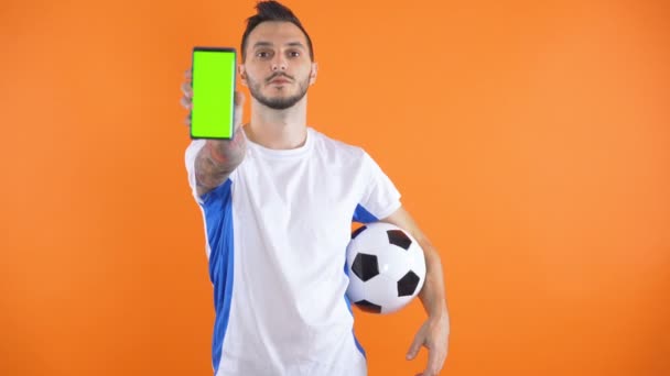 Футбольный болельщик В белой синей рубашке показать зеленый экран смартфона оранжевый фон  - Кадры, видео