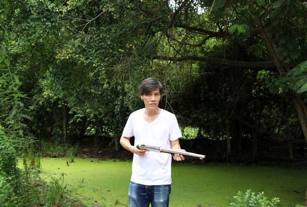 Střelec muž drží dlouhou zbraň v rukou a odbočil vlevo bokem s bílou košilí a džínem džín na zeleném pozadí stromu. - Fotografie, Obrázek