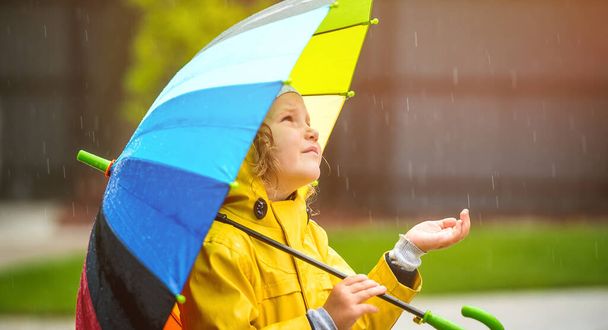 Lustiges kleines Mädchen, das im Garten im Herbstregen spielt. Kind trägt gelben wasserdichten Mantel und Stiefel mit buntem Regenschirm. Regenwetter. Kind fängt Tropfen auf - Foto, Bild