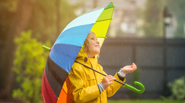 Funny little girl playing in the garden under the autumn rain. Kid wearing yellow waterproof coat and boots holding colorful umbrella. Rainy weather. Child is catching drops in sunny day - Photo, Image