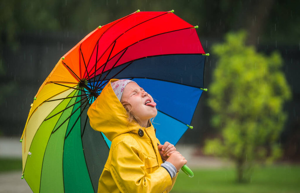 glückliches Kind, das Regentropfen im Park fängt. Lustiges kleines Mädchen, das im Herbstregen spielt. Kind trägt gelben wasserdichten Mantel und Stiefel mit buntem Regenschirm. - Foto, Bild