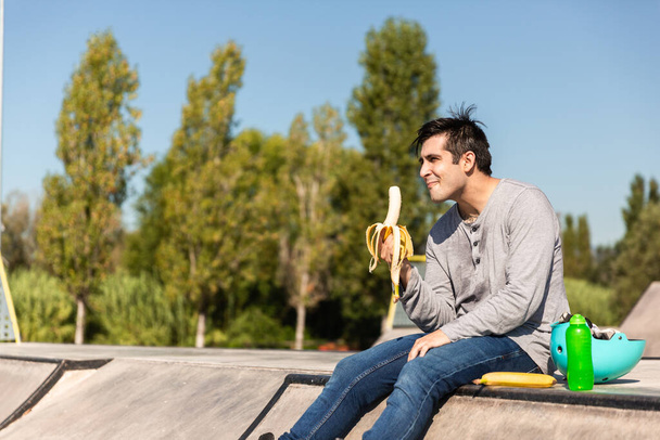 jongen met rolschaatsen eet een banaan zittend in een openbaar skatepark. Hij heeft een beschermhelm en een fles water op de zijkant. - Foto, afbeelding