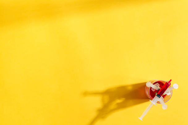Jeringas desechables estériles medicinales con vacuna roja o sangre en un vaso sobre un fondo amarillo con sombras largas, espacio para copiar. Vista superior. - Foto, Imagen
