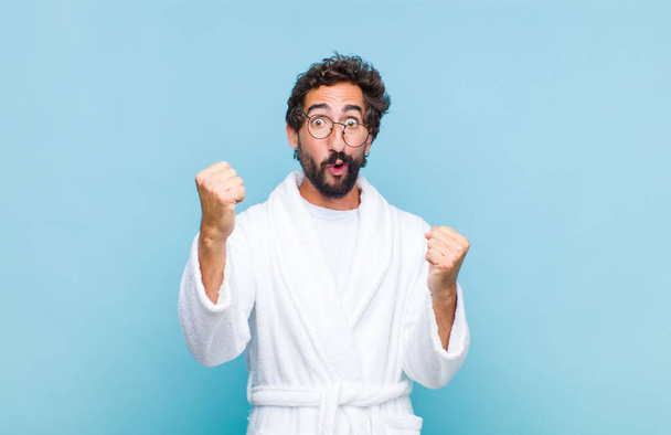 young bearded man wearing a bath robe celebrating an unbelievable success like a winner, looking excited and happy saying take that! - Φωτογραφία, εικόνα
