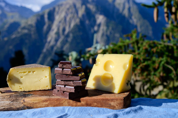 Νόστιμα ελβετικά τυριά και μαύρη καθαρή σοκολάτα, emmental, gruyere, appenzeller σερβίρεται εξωτερική με αλπικά βουνά κορυφές το καλοκαίρι στο παρασκήνιο - Φωτογραφία, εικόνα