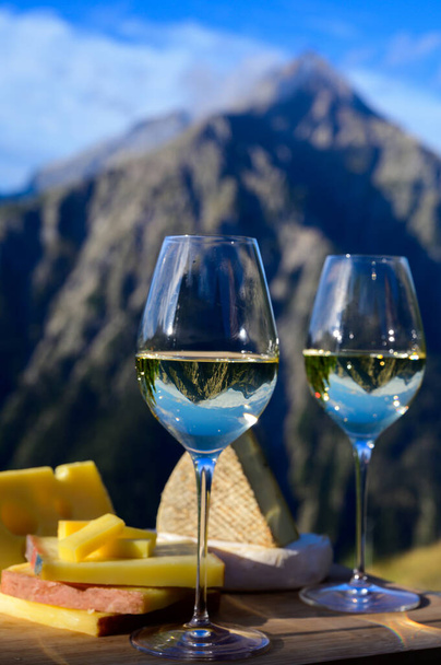 Lekkere kaas en wijn uit Savoie regio in Frankrijk, beaufort, abondance, emmental, tomme en reblochon de savoie kazen en glas witte wijn geserveerd buiten in de zomer met Alpenbergen pieken op de achtergrond - Foto, afbeelding