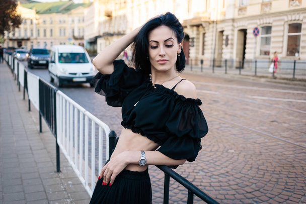 Ένα όμορφο κορίτσι με κοντά μαύρα μαλλιά σε μια μαύρη μπλούζα και φούστα στέκεται κοντά στο δρόμο, ακουμπώντας σε ένα φράχτη, το χέρι της στα μαλλιά και κοιτάζοντας προς τα κάτω. Στο κέντρο. Στην πόλη. Αυτοκίνητα στο παρασκήνιο. - Φωτογραφία, εικόνα