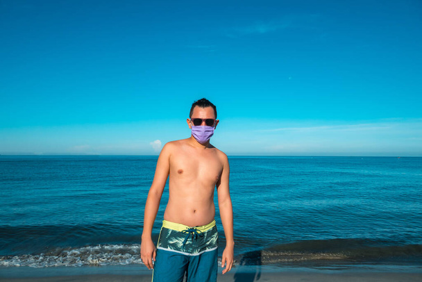 Hispanic man with sunglasses and a mask for the pandemic on the beach - 写真・画像