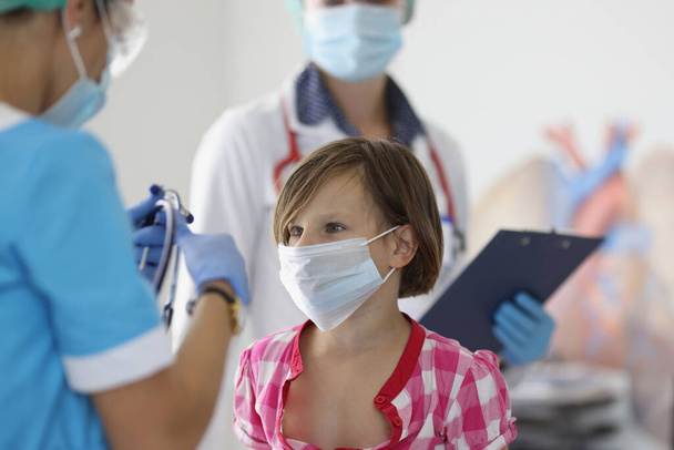 Klein meisje met beschermend medisch masker staat naast de dokter - Foto, afbeelding