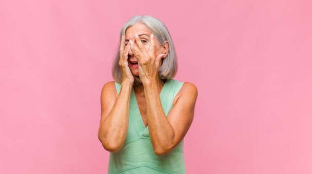 middle age pretty woman covering face with hands, peeking between fingers with surprised expression and looking to the side - 写真・画像