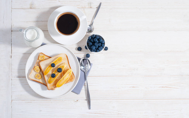 Πρωινό με μπανάνες και blueberry με φλιτζάνι καφέ και ψητές προπόσεις στο λευκό ξύλινο τραπέζι επίπεδη θέσει με χώρο αντίγραφο. Υγιεινό νόστιμο φαγητό το πρωί με νιφάδες βρώμης - Φωτογραφία, εικόνα
