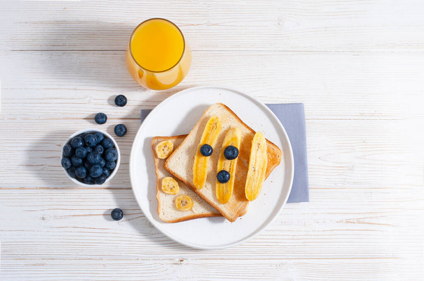 Breakfast with bananas and blueberry with orange juice and baked toasts on the white wooden table flat lay with copy space. Healthy tasty food at the morning with oat-flakes - Photo, image