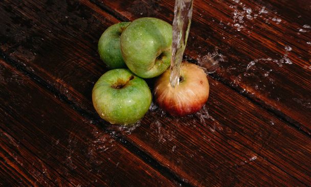 Des pommes rouges et vertes se trouvent sur la table de cuisine en acajou et de l'eau coule du haut. Des éclaboussures d'eau propre coulent sur les pommes fraîchement cueillies de la récolte d'automne. - Photo, image