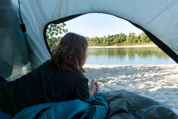 image rapprochée de la jeune femme se trouve dans la tente et regarde la belle rivière par la fenêtre ouverte. Camping dans de beaux endroits, mode de vie actif et sain. - Photo, image
