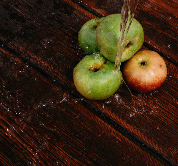 Rote und grüne Äpfel liegen auf dem mahagonifarbenen Küchentisch, von oben tropft Wasser. Spritzer sauberes Wasser ergießen sich über frisch gepflückte Äpfel der Herbsternte. - Foto, Bild