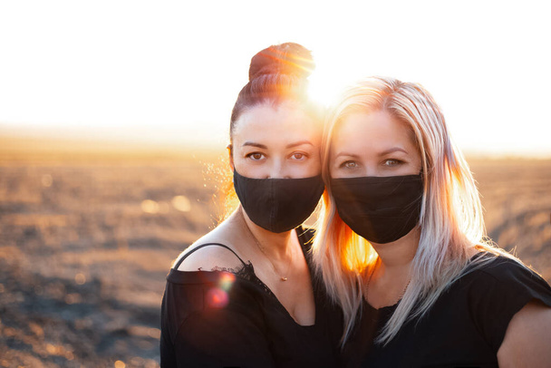 Outdoors portrait of two women, wearing respiratory face mask on the background of sunset. - Фото, изображение