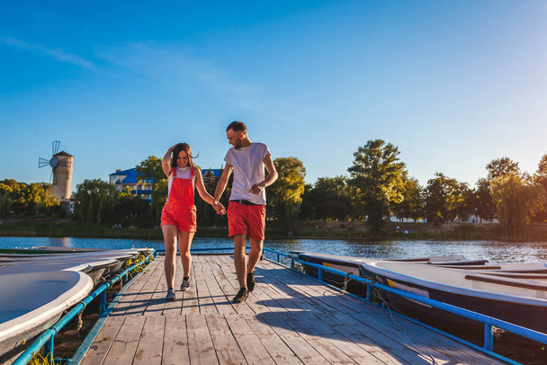 Young man and woman running along summer river dock with boats. Couple having fun at sunset holding hands. People relaxing outdoors - Foto, imagen