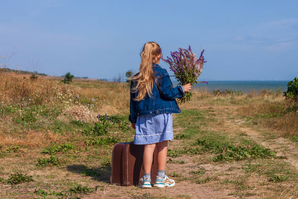 Little blonde girl in denim jacket, blue dress with vintage suitcase and flowers bouquet off-road with sea landscape. Stylish hitchhiker child with long hair on countryside trip. Kid walking outdoors. - Photo, Image