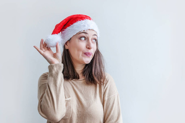 Krásná dívka v červené Santa Claus klobouk izolované na bílém pozadí vypadá šťastně a vzrušeně. Portrét mladé ženy, opravdové emoce. Veselé Vánoce a Nový rok. - Fotografie, Obrázek