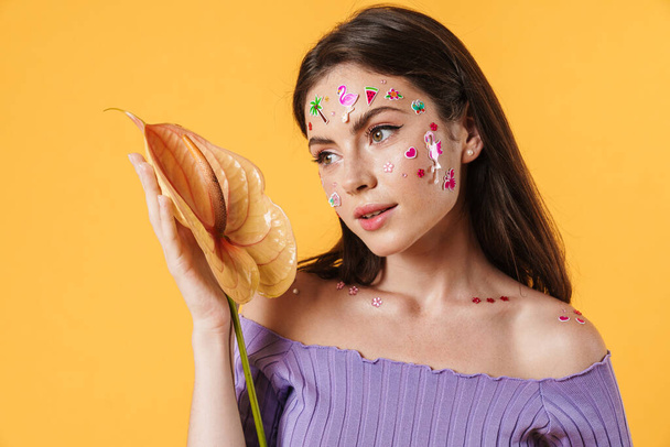 Image of young beautiful woman with stickers on face holding exotic flower isolated over yellow background - Φωτογραφία, εικόνα