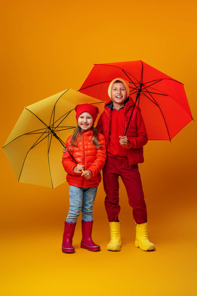 Happy emotional children, a boy and a girl, in autumn clothes, smiling and hugging standing under umbrellas. Children with orange and yellow umbrellas on a Colored yellow background. - Fotó, kép