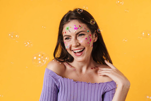 Image of young excited woman with stickers on face laughing at camera isolated over yellow background - Foto, afbeelding