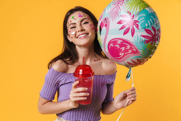 Image of laughing woman with stickers on face holding balloon and drinking cocktail isolated over yellow wall - Foto, imagen