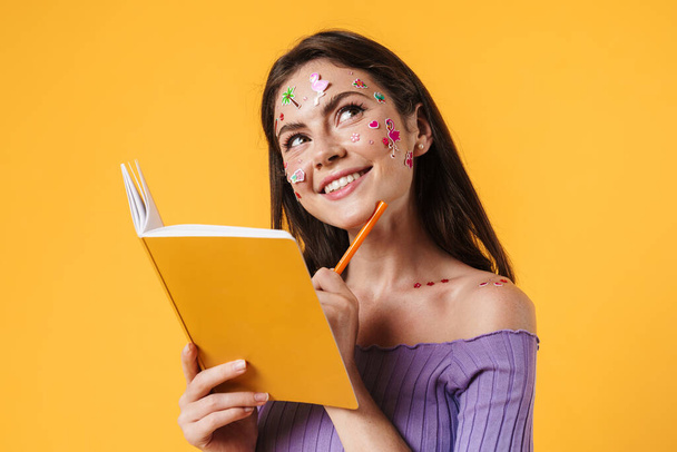 Image of young smiling woman with stickers on face writing in exercise book isolated over yellow background - Foto, immagini