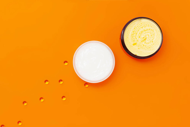 Две банки с различными кремами на оранжевом фоне рядом с которыми находятся капсулы с витамином Е. - Фото, изображение