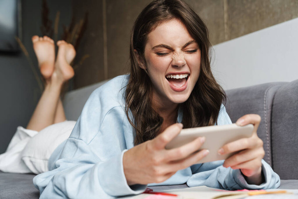 Image of excited nice woman playing online game on mobile phone while lying on sofa at home - Valokuva, kuva