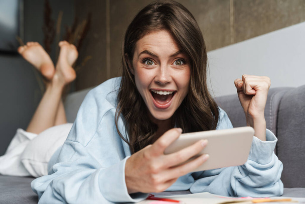 Imagen de mujer emocionada haciendo gesto ganador y jugando juego en línea en el teléfono celular mientras está acostado en el sofá - Foto, imagen
