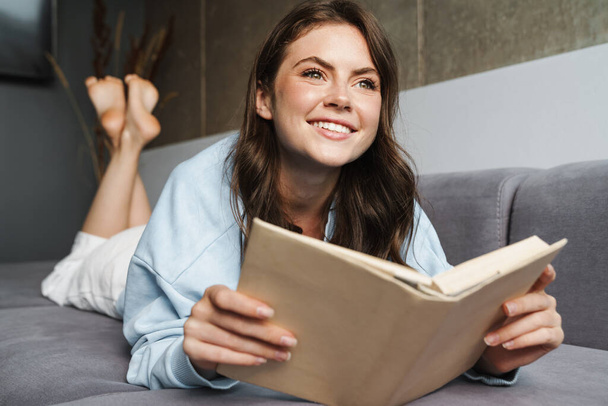 Image of cheerful nice woman smiling and reading book while lying on sofa at home - Фото, зображення