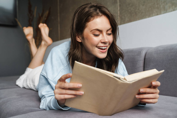 Image of cheerful nice woman smiling and reading book while lying on sofa at home - Foto, afbeelding