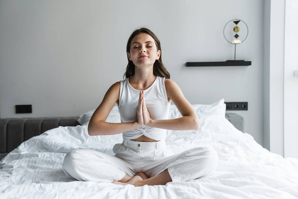Image of beautiful calm woman meditating and holding palms together while sitting on bed at home - Foto, Imagen