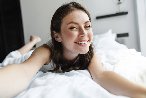 Image of charming happy woman smiling and taking selfie photo while lying in bed at home - Fotoğraf, Görsel